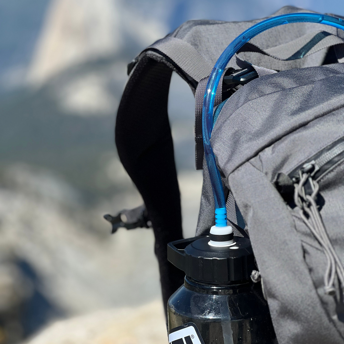 A close-up of a gray hiking backpack with a blue drinking tube and a black Yeti water bottle attached, set against a blurred mountain landscape.